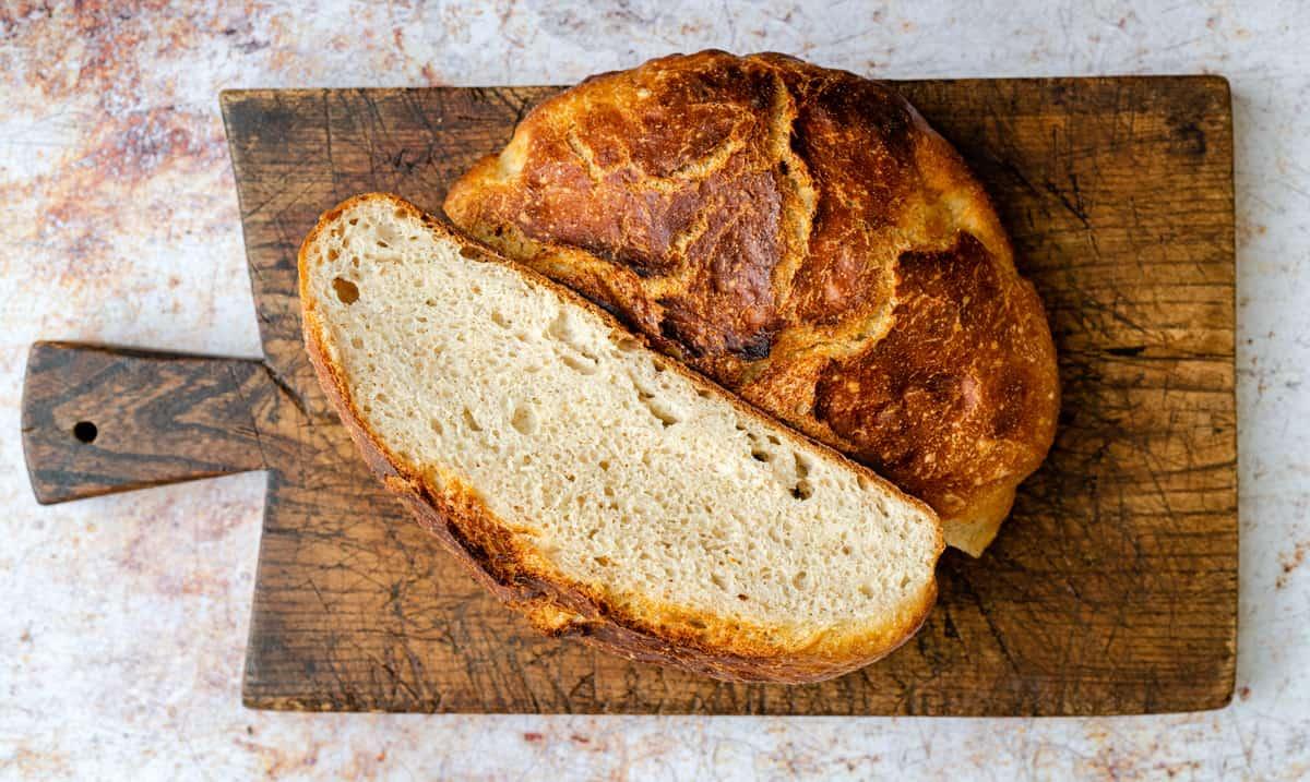 A round loaf of rustic sourdough bread with one thick slice cut, sits on a weathered wooden cutting board against a textured background.