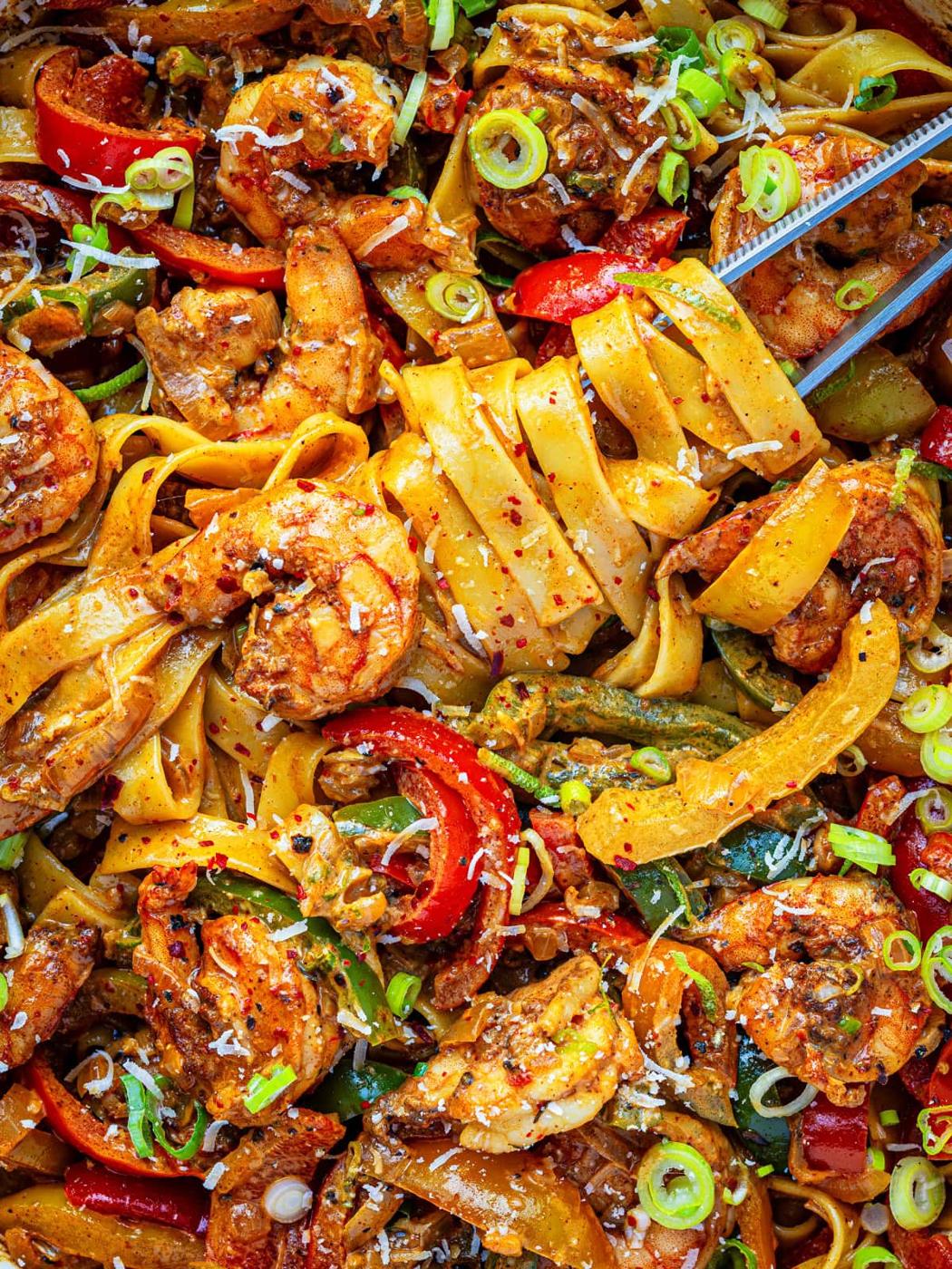 A close-up of rasta pasta with jerk shrimp with wide noodles, shrimp, sliced red peppers, green onions, and a sprinkle of red pepper flakes, garnished with grated cheese and herbs.