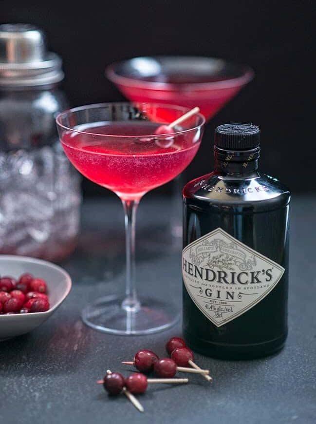 A Red Queen cocktail in a coupe glass garnished with cranberries, next to a bottle of Hendrick’s gin, a cocktail shaker and a bowl of fresh cranberries.