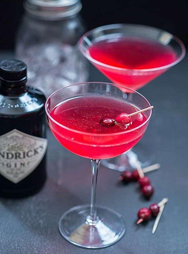 Two ruby red Red Queen cocktails in coupe glasses garnished with cranberries, with a bottle of gin behind and extra cranberries on cocktail picks.