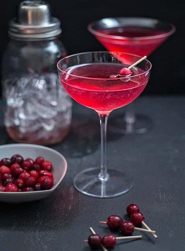 A Red Queen gin cocktail in a coupe glass garnished with cranberries, with a bowl of fresh cranberries, a cocktail shaker and cranberries on cocktail picks.