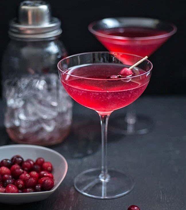 A cranberry and elderflower gin cocktail in a coupe glass with a cranberry garnish, with a second cocktail and a shaker of ice in the background.