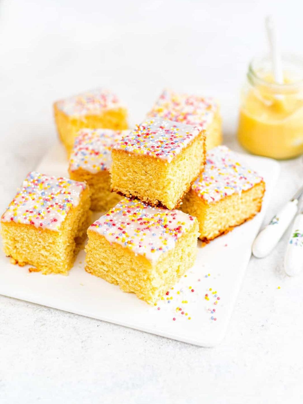 Slices of classic school cake, with colorful sprinkles are arranged on a white surface. A jar with a custard and two forks are in the background.