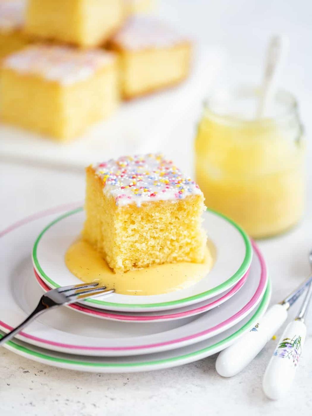 A slice of vanilla school cake with white icing and colorful sprinkles sits on stacked plates. A jar of custard with a spoon is in the background. Two forks rest beside the plates on a light surface, ready for a delightful treat.