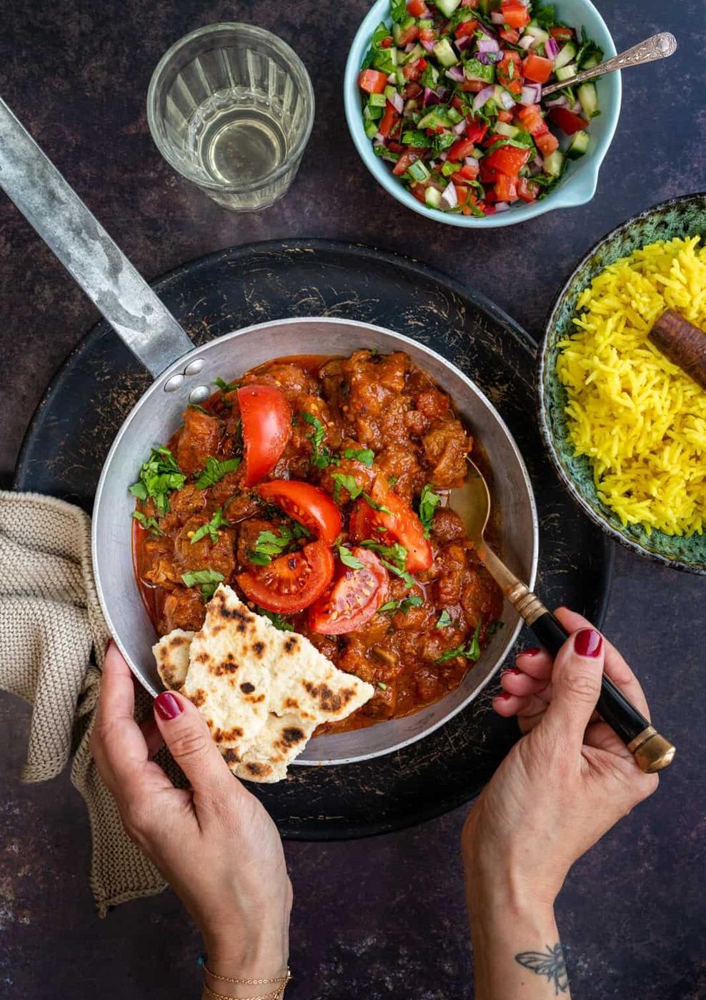 A person holds naan bread and a fork, about to eat a pan of lamb rogan josh, a rich tomato-based curry garnished with cilantro and tomato. Nearby are a bowl of yellow rice, salad, and a glass of water on a dark tabletop.