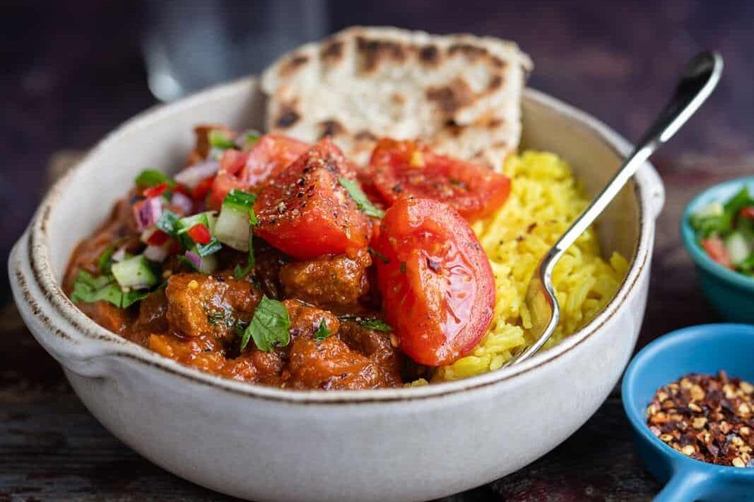 A bowl of curry with chunks of meat, yellow rice, sliced tomatoes, and fresh herbs, served with flatbread. A spoon rests in the bowl, and small bowls of chili flakes and chopped vegetables are beside it.