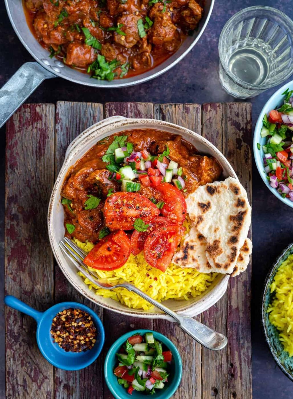 Lamb rogan josh in a bowl with rice and kachumber salad on a set table