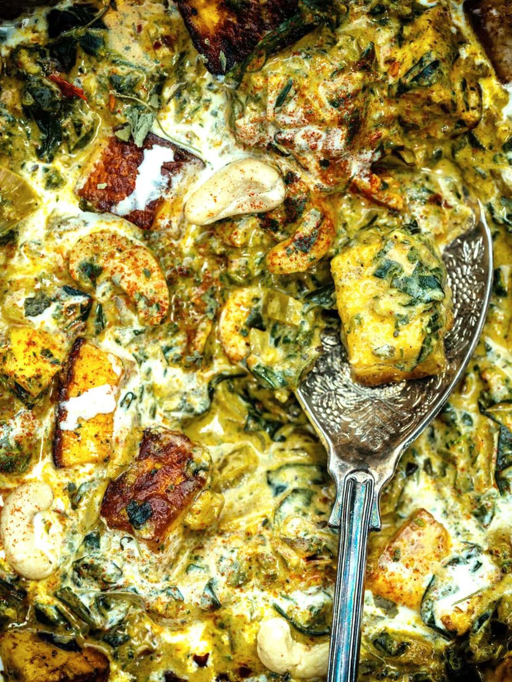 Close-up of a creamy Saag Paneer curry with paneer cubes, cashews, spinach, and spices, with a silver serving spoon resting in the dish. The sauce appears rich and well-seasoned.