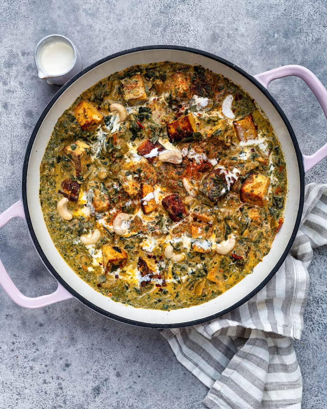 A round pan with handles holds a creamy spinach curry, or saag paneer, topped with paneer cubes, cashews, and a drizzle of cream. A small jug of cream and a striped cloth sit beside the pan on a grey surface.