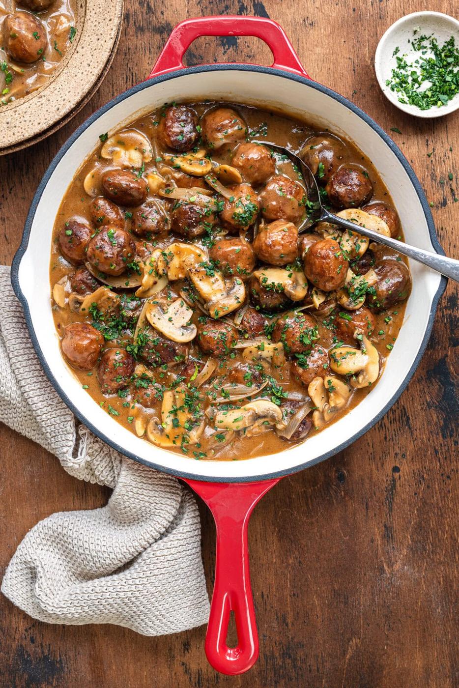 Salisbury steak meatballs simmering in a rich mushroom and onion gravy in a red skillet, garnished with chopped parsley, with a spoon resting in the dish.