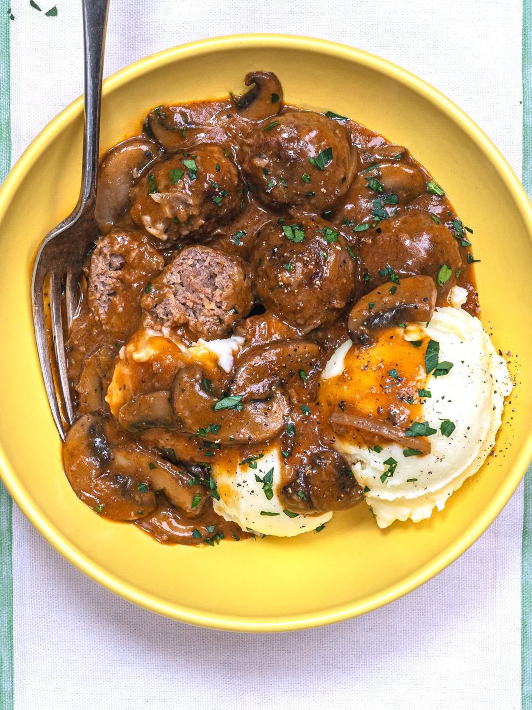 A yellow plate with Salisbury steak meatballs in brown mushroom gravy, served alongside mashed potatoes with melted butter and herbs, and a fork on the side.