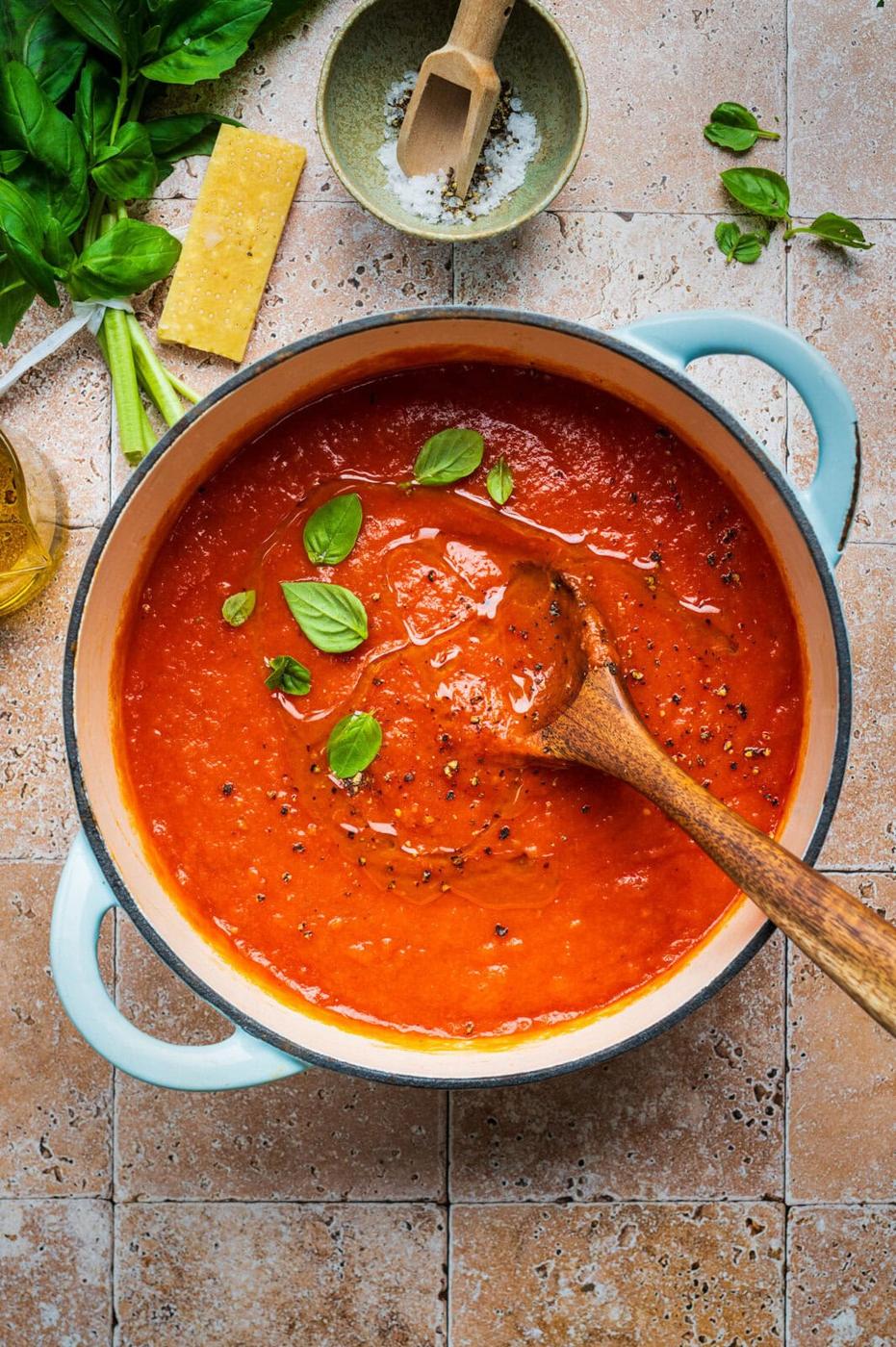A pot of San Marzano tomato sauce garnished with basil leaves sits on a tiled surface. A wooden spoon rests inside the pot. Fresh basil, a wedge of cheese, salt, and olive oil are arranged nearby.
