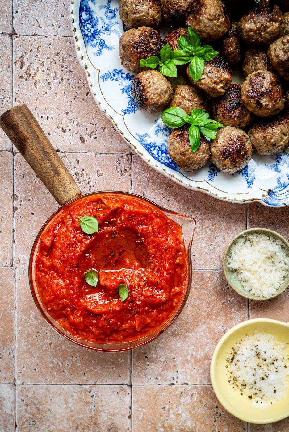 A bowl of chunky tomato sauce garnished with basil sits beside a plate of meatballs topped with fresh basil, with small bowls of grated cheese and olive oil on a tiled surface.
