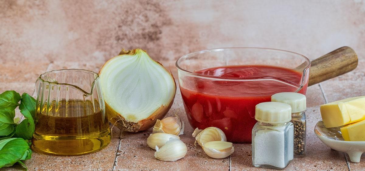 A glass jug of olive oil, half a white onion, a glass bowl of crushed tomatoes, garlic cloves, basil leaves, salt and pepper shakers, and pieces of butter are arranged on a tiled surface.
