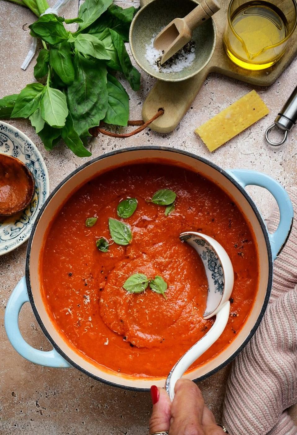 A hand ladles rich tomato soup from a blue pot, garnished with fresh basil leaves. Surrounding the pot are basil, olive oil, salt, a cheese wedge, and a small plate with more sauce.