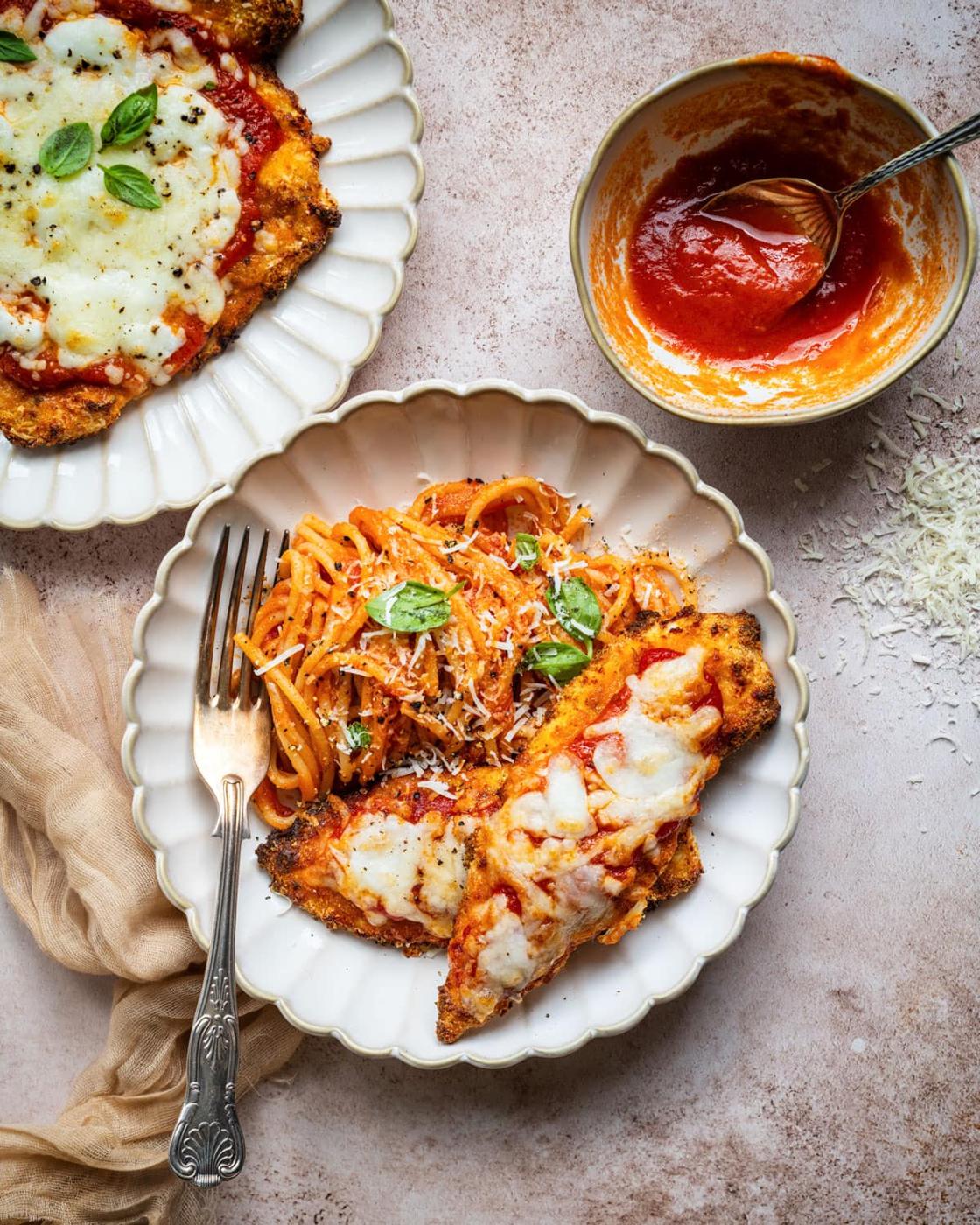 A plate with spaghetti topped with tomato sauce and grated cheese, served alongside chicken parmesan with melted cheese. Another plate with chicken parmesan and a small bowl of tomato sauce are nearby.