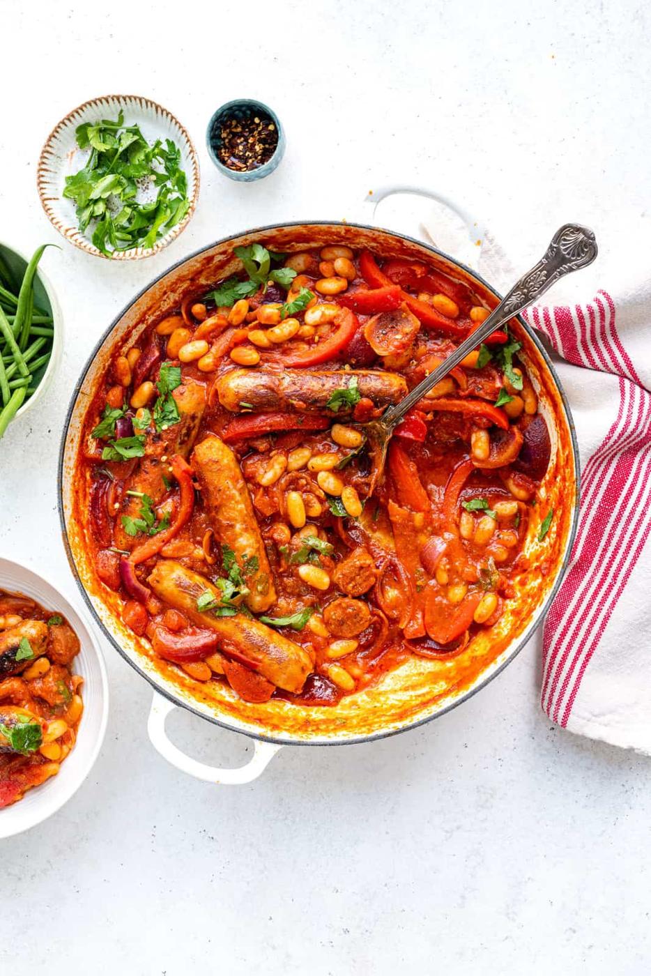 A white pot filled with Easy Sausage Casserole—sausage and bean stew in a rich tomato sauce, topped with chopped herbs. A spoon rests in the pot, with a red-striped towel, small bowl of herbs, and bowl of green beans nearby.