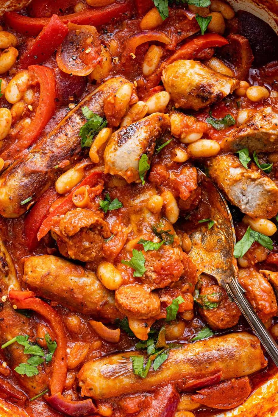 A close-up of a hearty Sausage and Bean Casserole with sliced sausages, white beans, tomatoes, red bell peppers, and chopped herbs simmered in a rich tomato sauce, with a spoon partially visible.