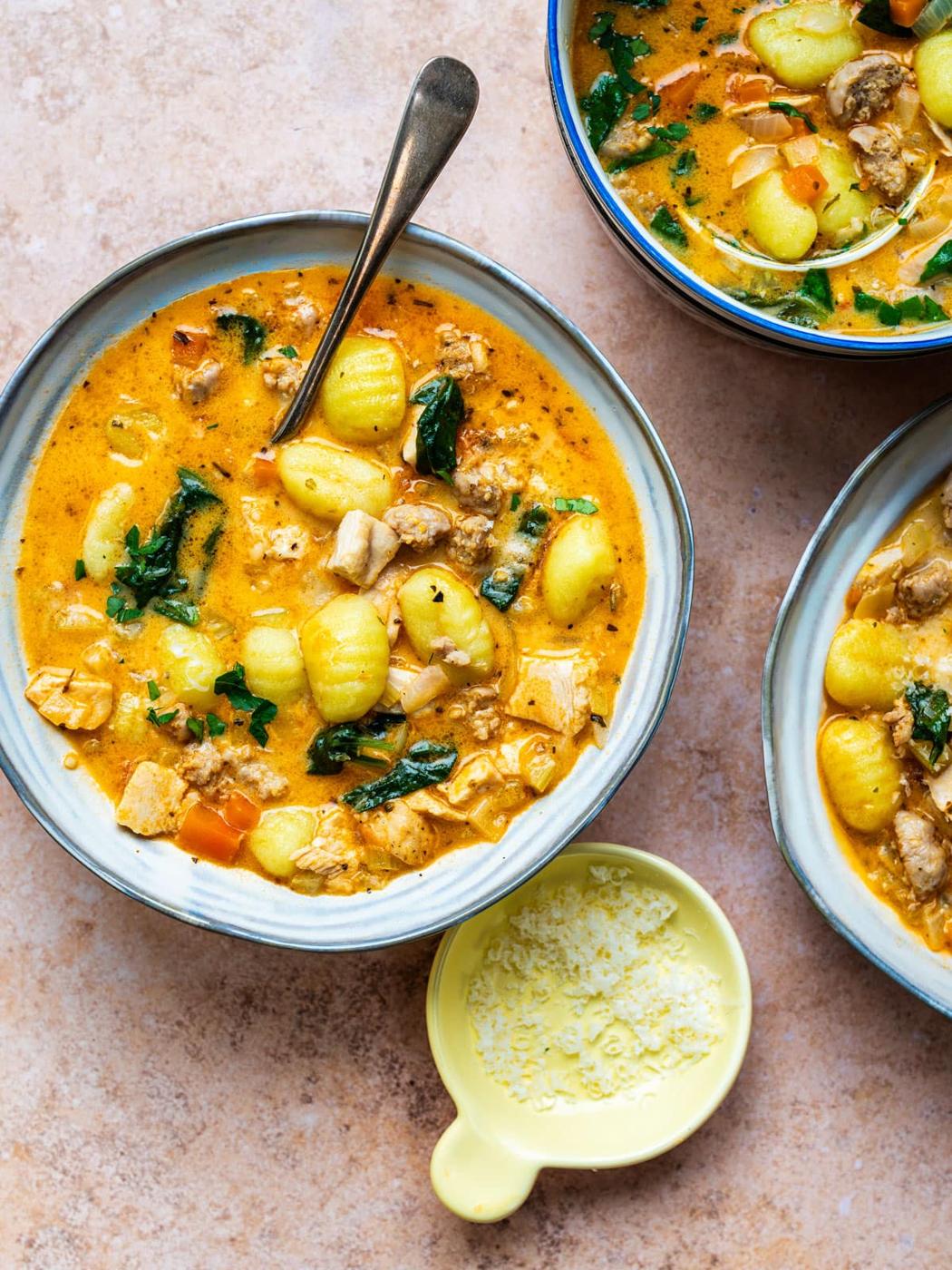 Three bowls of creamy gnocchi soup with chicken, spinach, carrots, and herbs, garnished with fresh greens. Try substituting Italian sausage or leftover turkey for a delicious twist. A small yellow dish of grated cheese sits nearby.