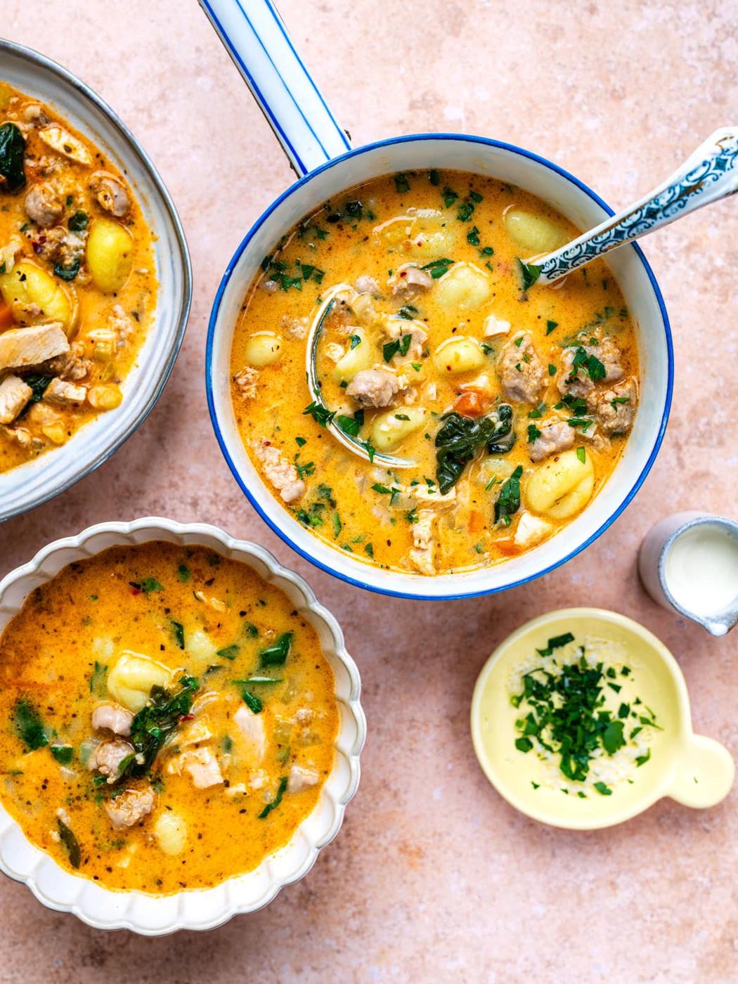 A pot and two bowls of Creamy Sausage Gnocchi Soup with Leftover Turkey, spinach, and herbs sit on a light countertop. Fresh parsley and a small jug of cream are nearby, ready for serving.