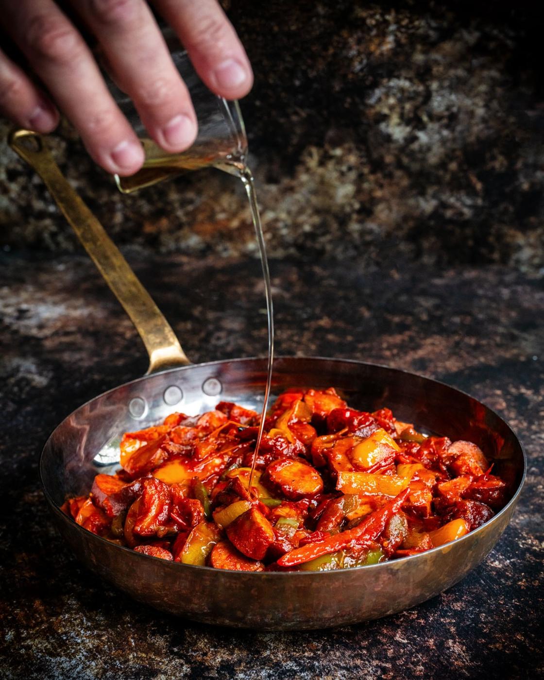 A hand pours ouzo from a glass into a copper pan filled with a cooked mixture of sliced sausage and peppers in a rich tomato sauce