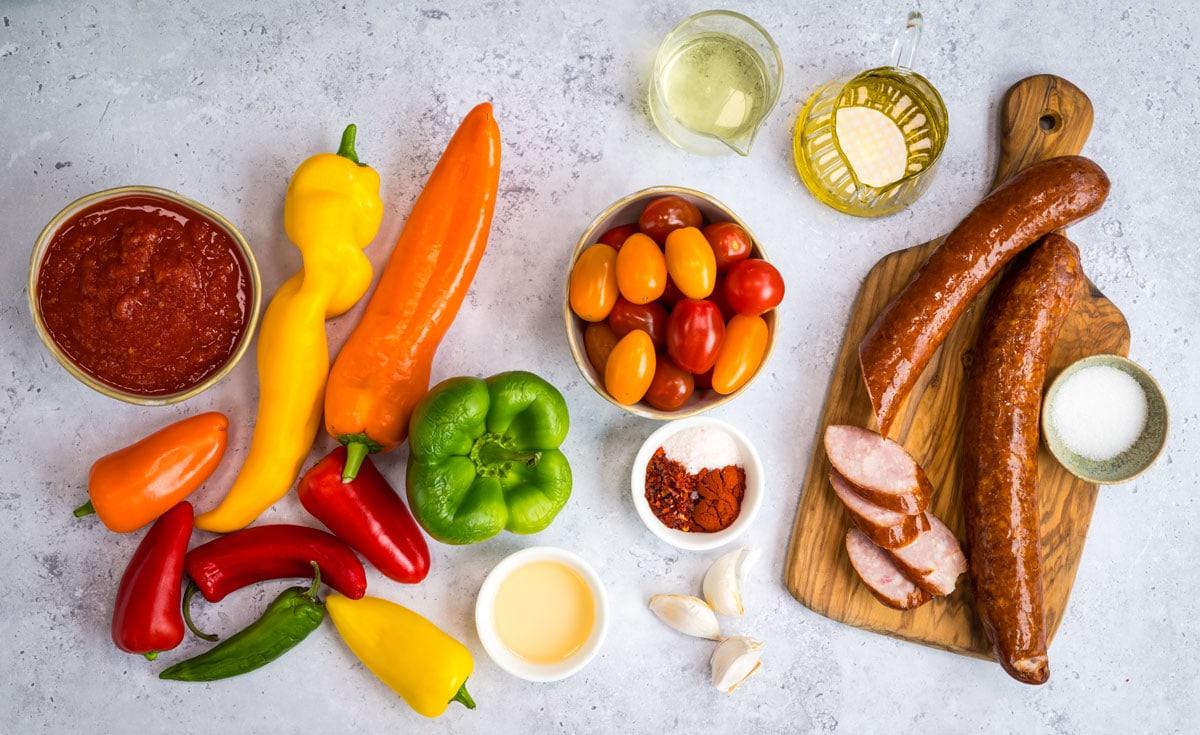 Assorted colorful peppers, tomatoes, green bell pepper, garlic, sausage, tomato sauce, oil, vinegar, spices, and salt arranged on a gray surface and wooden board.