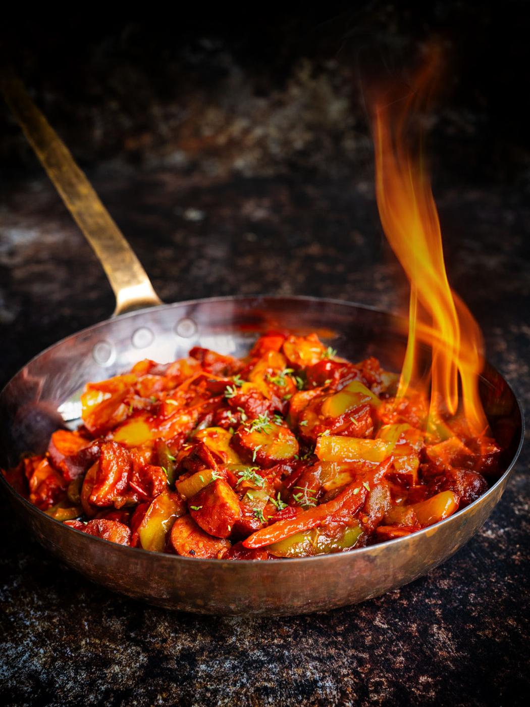 sausage and pepper stew in a copper pan, flames coming out of the dish as it is being flambéed
