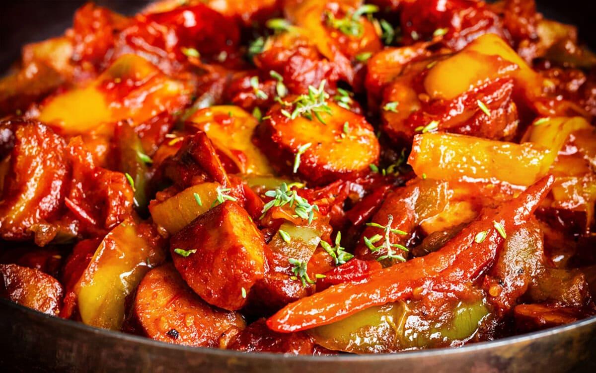 Close-up of a dish featuring sliced sausage, bell peppers, and onions cooked in a rich red tomato sauce, garnished with fresh herbs. The food is vibrant and appears hot and flavorful.