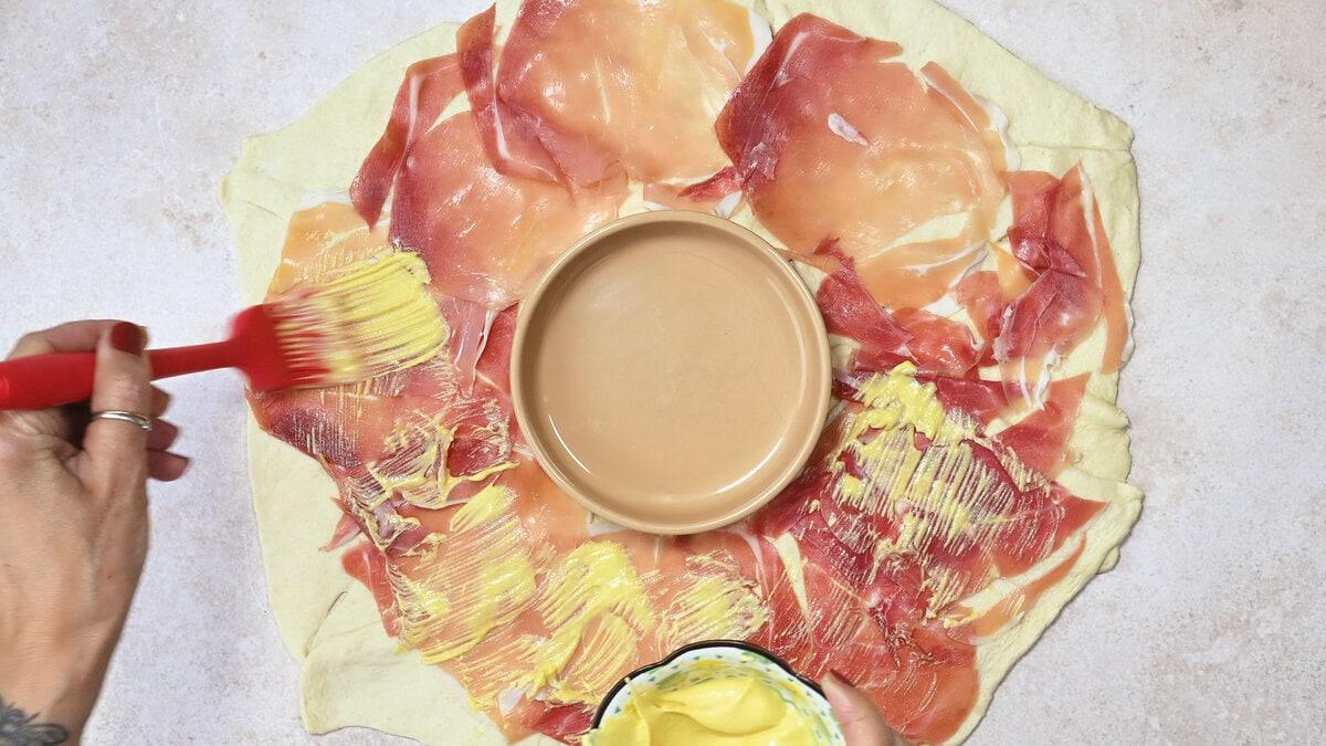 crescent roll pastry arranged in a circle topped with prosciutto slices and brushed with mustard