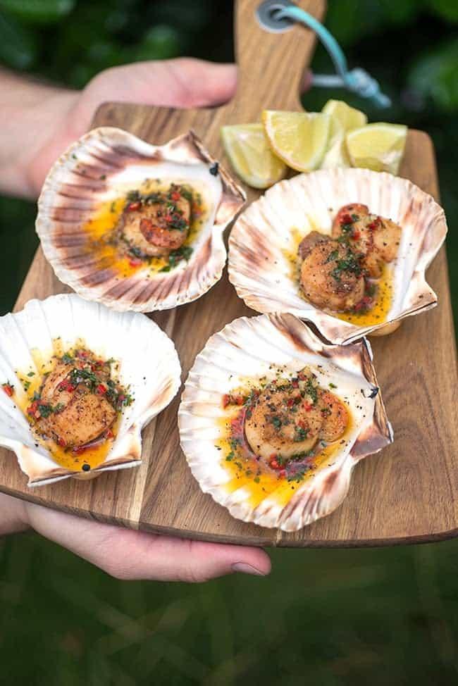 A wooden board holds four scallop shells filled with cooked scallops in a seasoned sauce, garnished with herbs and spices, alongside several lime wedges.