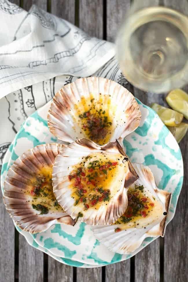 A plate of cooked scallops in shells topped with herbs and sauce, set on a blue and white patterned plate; a drink and lime wedges are next to the plate on a wooden table.