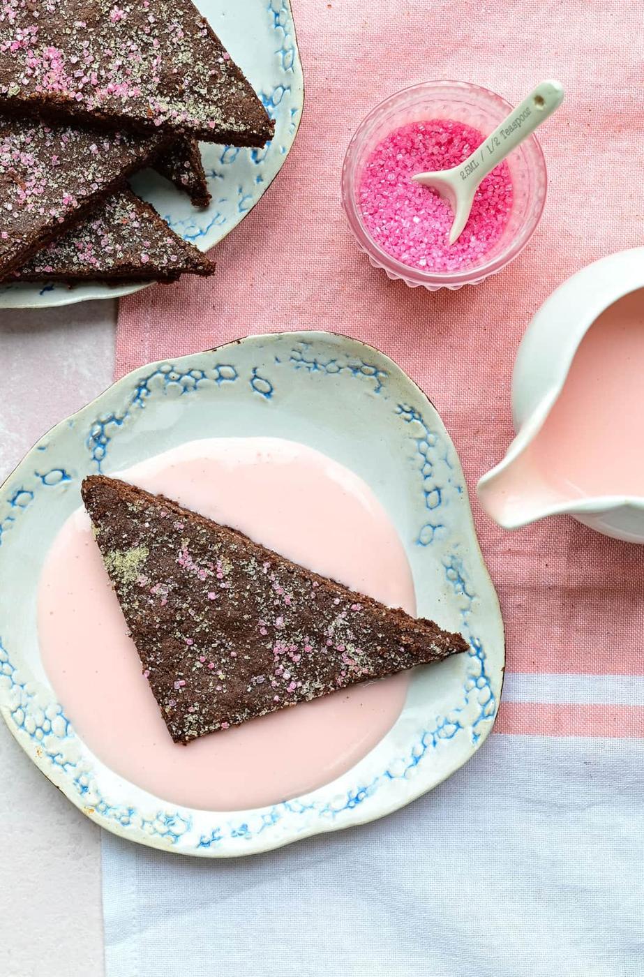 A triangular piece of chocolate concrete dessert with pink sprinkles sits on a floral plate, surrounded by pink custard. Nearby, a small bowl of pink sprinkles and a pitcher of vibrant pink sauce rest on a pink and white cloth.