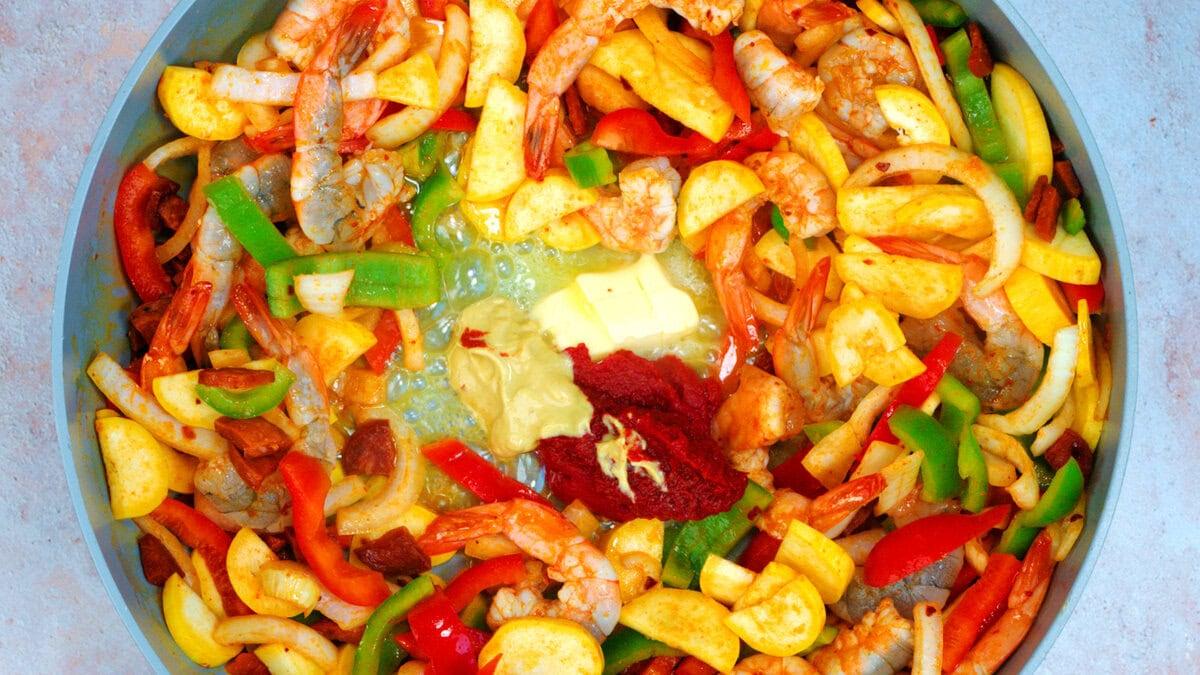 Colorful pan filled with raw shrimp, sliced bell peppers, onions, squash, chorizo, butter, mustard, and tomato paste, ready to be cooked.