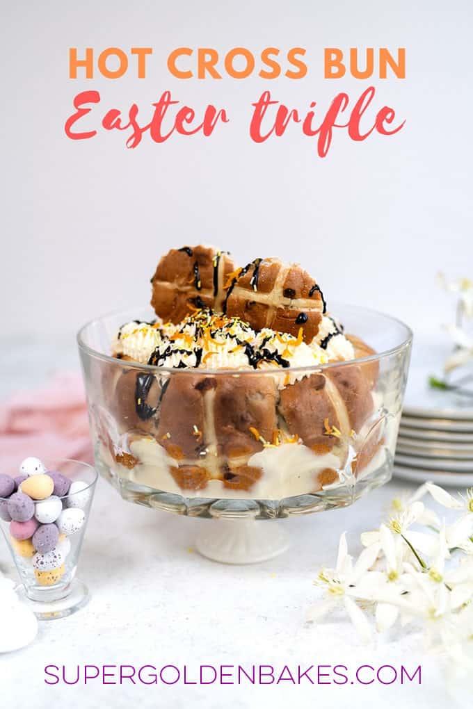 A glass bowl of Easter trifle crowned with hot cross buns, whipped cream, and a chocolate drizzle. Resting on a white surface, it’s surrounded by flowers and decorative eggs. Above it, the words Hot Cross Bun Easter Trifle shine brightly.
