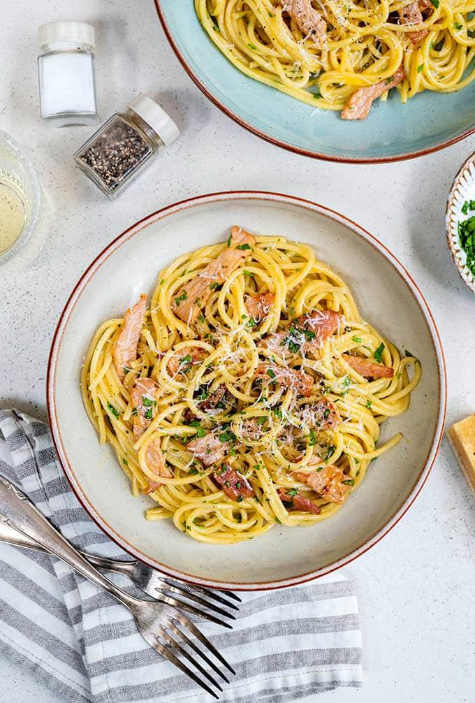 A bowl of spaghetti carbonara topped with bacon, grated Parmesan and parsley, with another plate of pasta, salt and pepper shakers, and a striped napkin with forks nearby on a light surface.