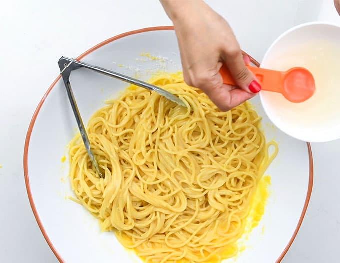 A hand with red nail polish pours liquid from a small white bowl with an orange spoon into a large bowl of yellow pasta, preparing an authentic Carbonara recipe, with metal tongs resting on the side.