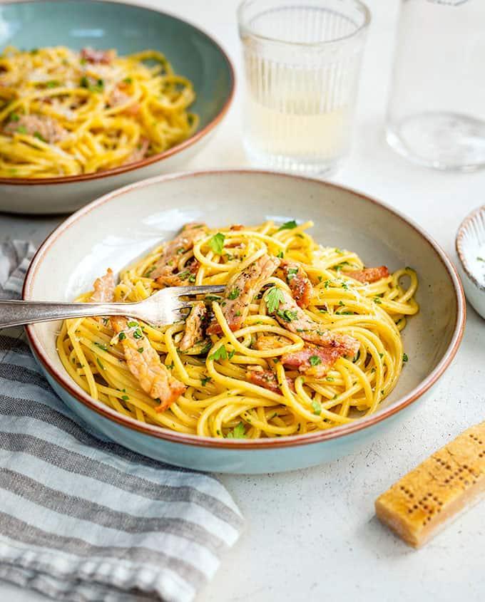 A bowl of authentic Carbonara with bacon, grated cheese, and parsley is served with a fork. A glass of water, another pasta bowl, a striped napkin, and a wedge of cheese rest nearby on a light table.