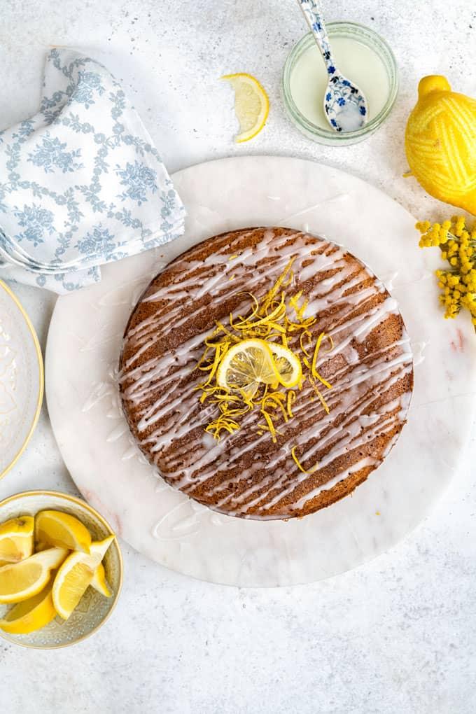 A round Slimming World Lemon Drizzle Cake with white icing, garnished with a lemon slice and zest. Its elegantly set among a blue and white napkin, a lemon juicer, a small bowl with a spoon, and a plate of lemon wedges on a light surface.