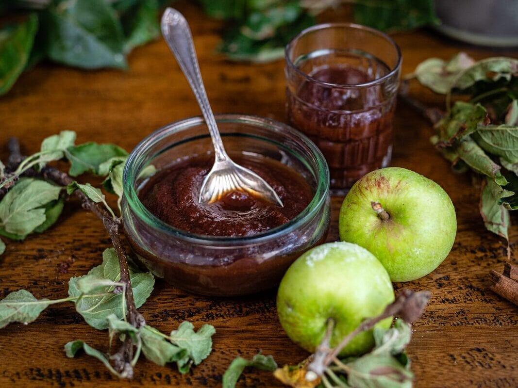 A glass bowl of apple butter with a spoon, a glass of juice, green apples, and apple branches on a wooden table, with a bowl of apples in the background.