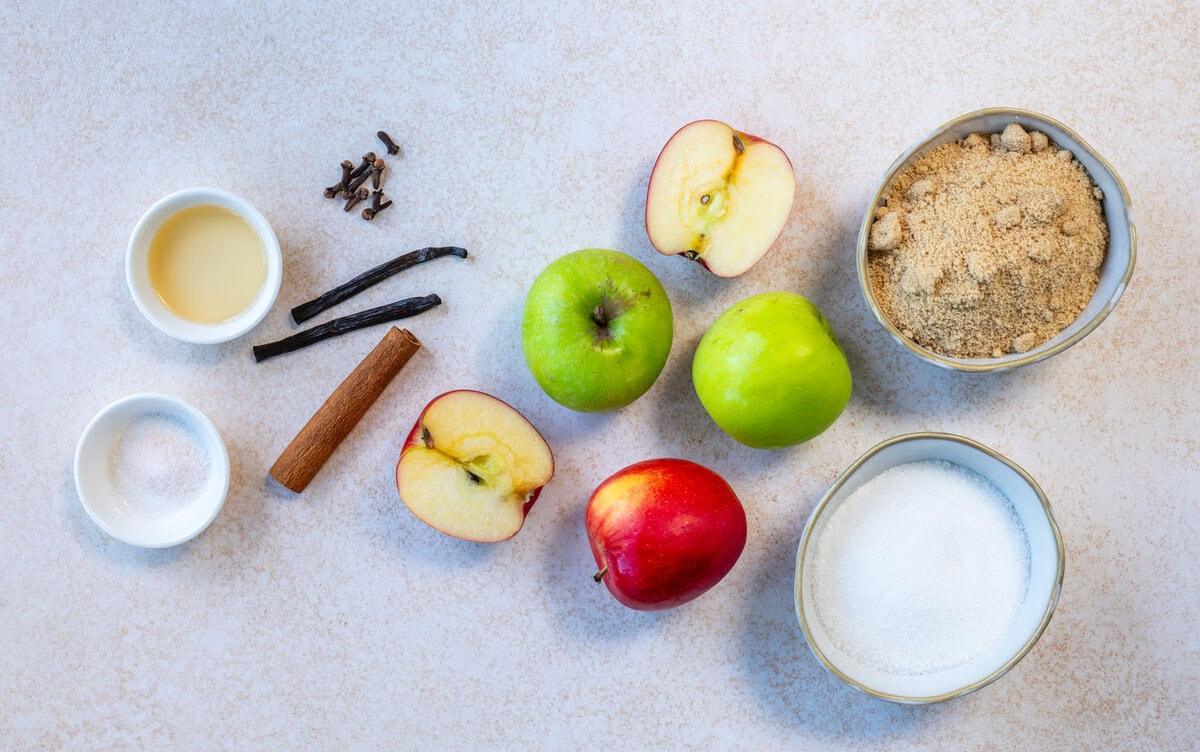 Three whole apples, two apple halves, a bowl of brown sugar, a bowl of white sugar, a cinnamon stick, vanilla beans, cloves, salt, and a small bowl of liquid are arranged on a light surface.