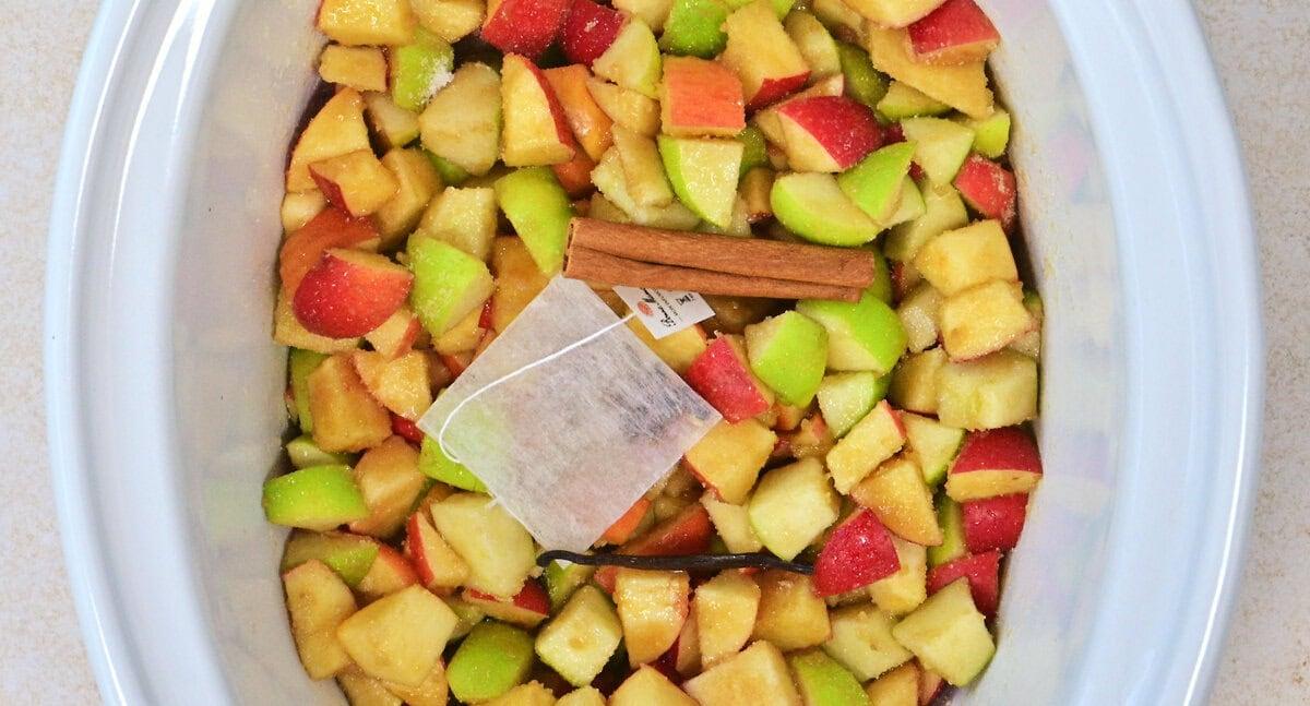 Chopped red and green apples in a slow cooker with a tea bag, a vanilla bean, two cinnamon sticks, and sprinkled sugar on top.