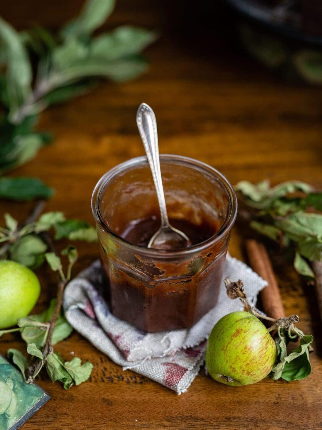 A glass jar filled with apple butter and a spoon inside is placed on a cloth napkin. Fresh green apples, cinnamon sticks, and leafy apple branches are arranged around it on a wooden table.