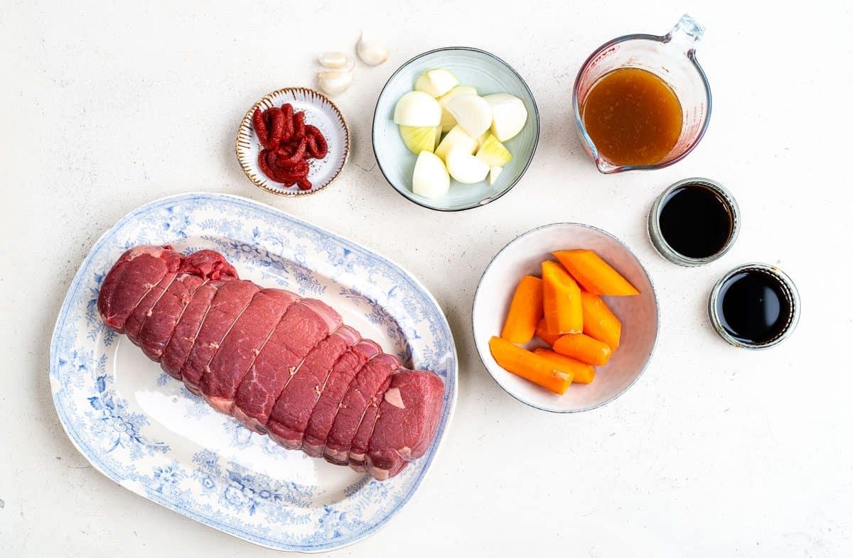 raw large beef joint on a platter surrounded but sliced carrots, onions, garlic, stock, tomato paste