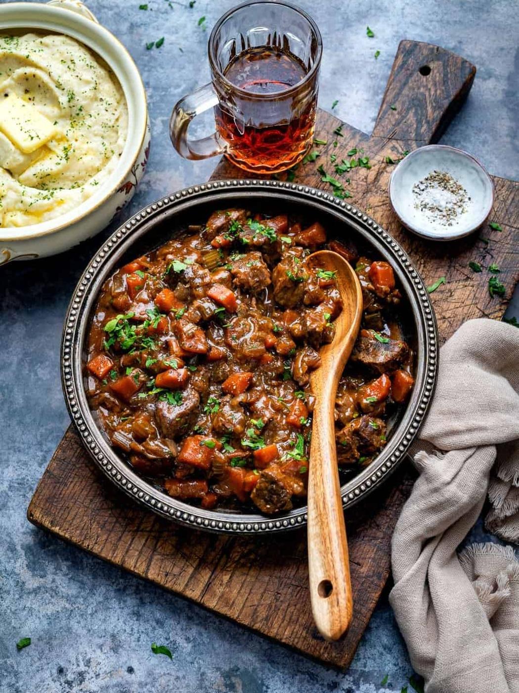 A bowl of hearty beef and ale stew with carrots and herbs sits on a wooden board, next to mashed potatoes, a dark drink, pepper, and a beige napkin. A wooden spoon rests in the slow cooker stew.