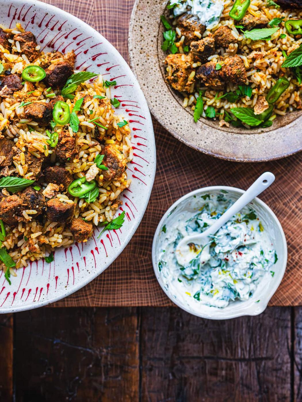A large plate and a bowl of slow cooker beef biryani topped with green chili slices and fresh mint leaves. Nearby is a small bowl of creamy sauce with herbs and a spoon. Everything is set on a wooden surface.