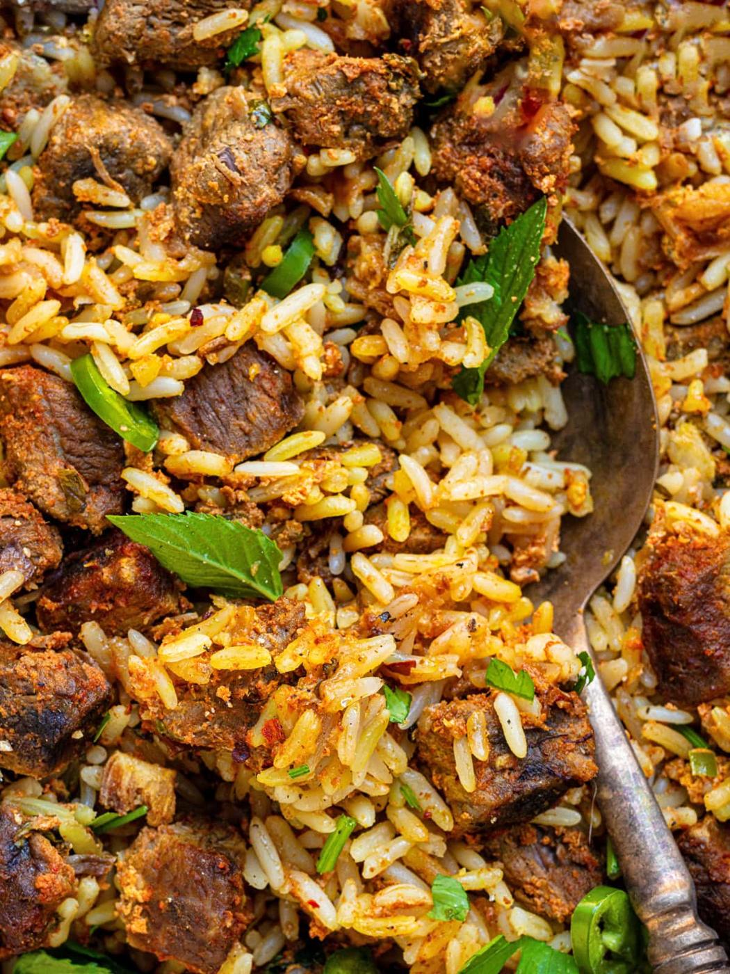 Close-up of a flavorful crockpot beef biryani with chunks of spiced meat, garnished with fresh green herbs. A spoon is partially visible, scooping up the rice and meat mixture.