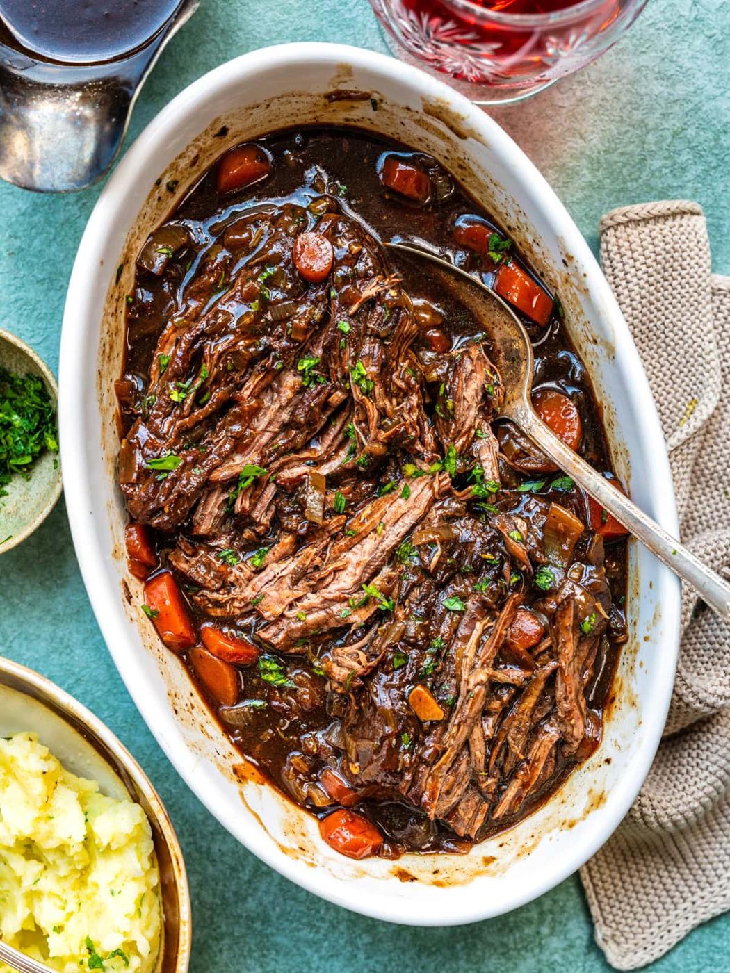 A white oval dish filled with shredded crockpot braised steak in rich brown gravy with carrots and herbs, next to a bowl of mashed potatoes, chopped parsley, and a glass of red wine. A spoon rests in the roast.