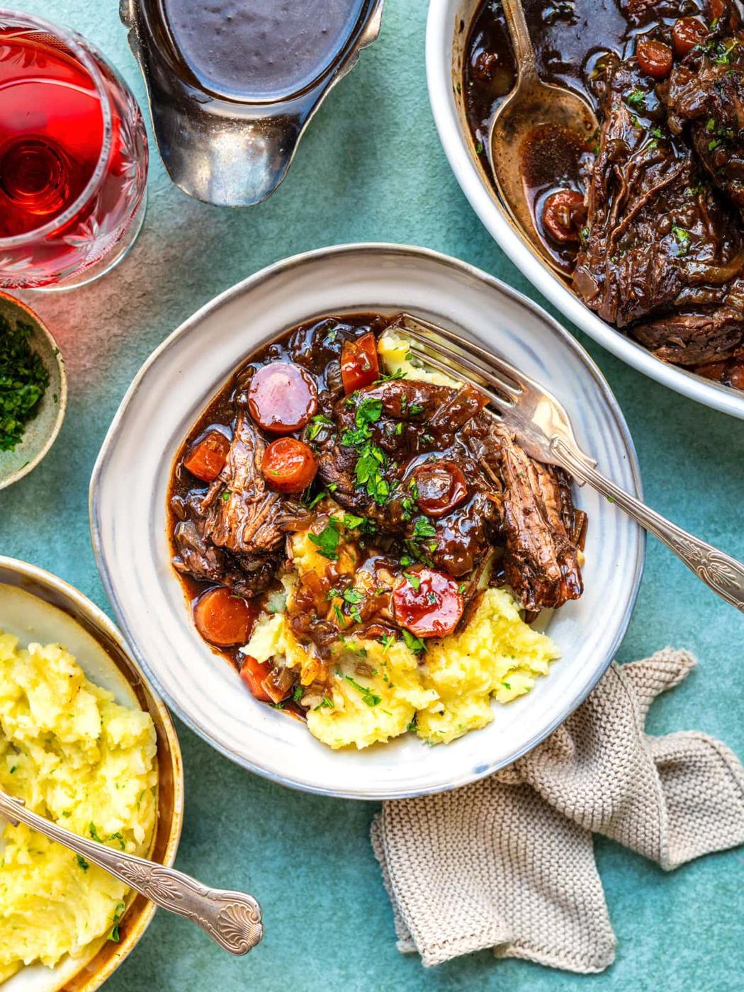 A bowl of mashed potatoes topped with braised steak, carrots, and rich brown gravy sits on a table. Nearby are a bowl of extra mashed potatoes, a dish of braised beef, a glass of red wine, and a sauce boat.