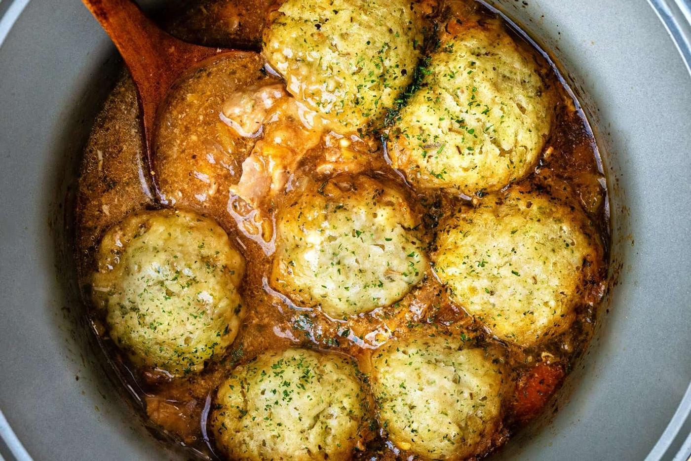 A pot of Slow Cooker Chicken and Dumplings with golden-brown dumplings simmering on top, garnished with herbs. A wooden spoon stirs the thick, flavorful broth surrounding the dumplings.