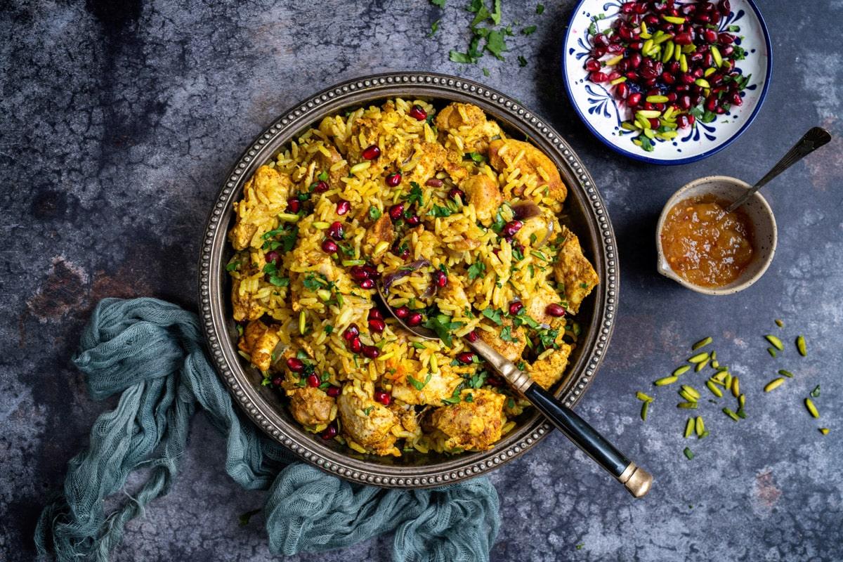 Chicken Biryani in a bowl with a side bowl of chutney and plate of pomegranate seeds and sliced pistachios